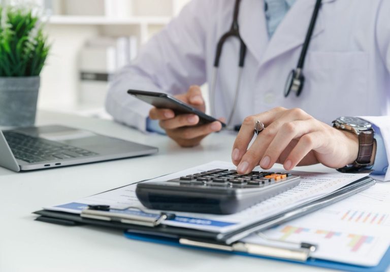 Um médico de jaleco usa calculadora e celular, com laptop e papéis sobre a mesa. Curso: Obrigações Econômico-Financeiras ANS.