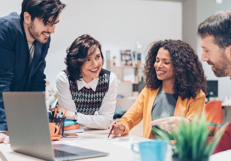 Quatro pessoas discutem ideias em um escritório com laptop, papéis e planta sobre a mesa. Curso: Pensar Fora da Caixa.