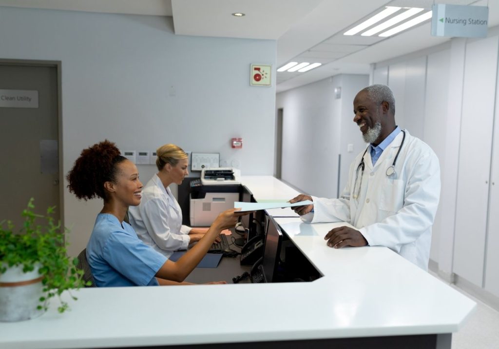 Médico e recepcionistas em balcão, com documentos e planta ao lado. Curso: Resultados e Melhorias.