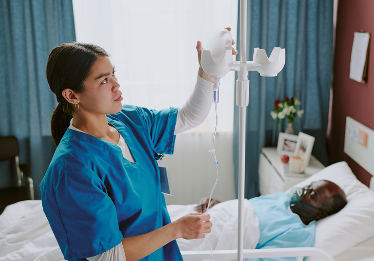 Mulher formada no curso Técnico em Enfermagem está ajustando soro de paciente internado em cama de hospital, com flores e fotos ao lado em um quarto acolhedor.