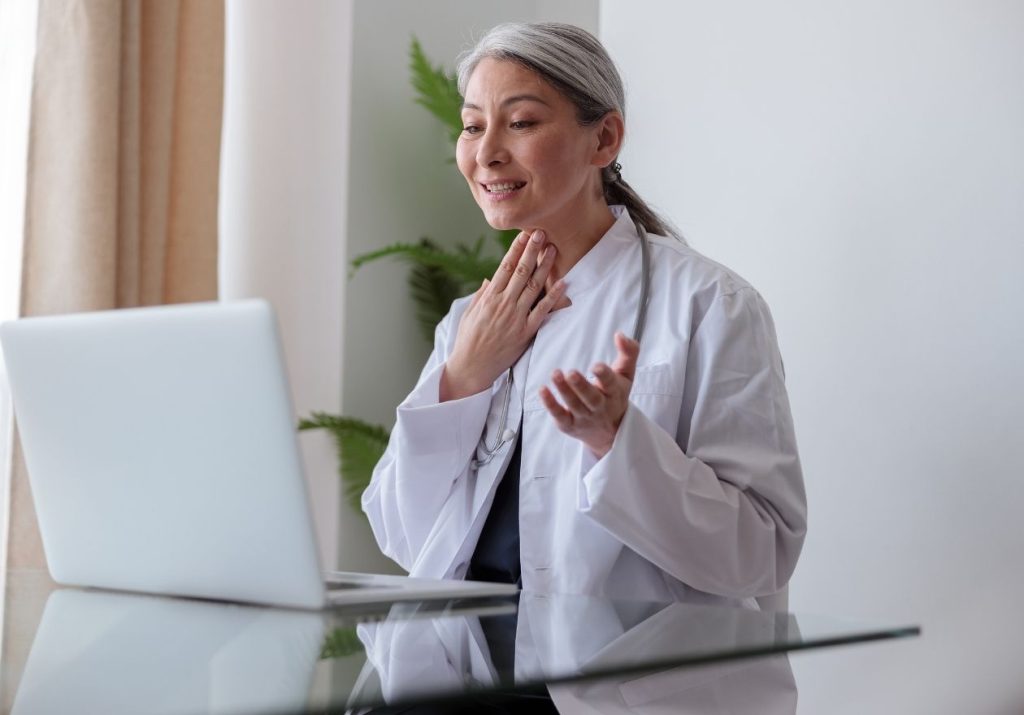 Médica em jaleco branco, gesticulando na frente de um laptop, em um ambiente claro com planta ao fundo. Curso: Telemedicina.